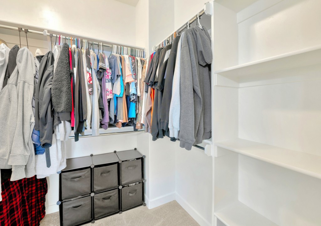 Small walk-in closet with hanging sweaters, shirts and blouse on the metal rods. Closet with linen drawers on below the clothes on the left and a view of an empty vertical framed shelves on the right.